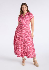 Pink maxi dress with a geometric pattern in cream. Features short sleeves, a wrap design, and a flowing silhouette. Worn with wedge sandals.