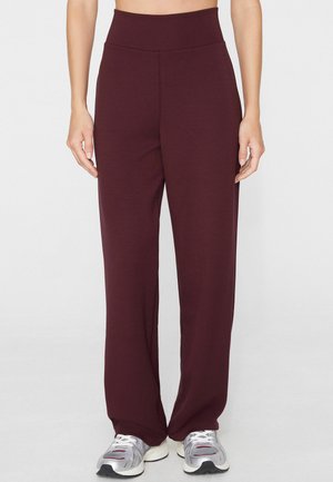Persona che indossa pantaloni a vita alta, ampi, di colore bordeaux scuro, abbinati a scarpe da ginnastica argento e bianche, in piedi davanti a uno sfondo chiaro uniforme.