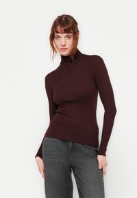Rollkragenpullover aus dunkelburgunderfarbenem Rippstrickstoff, taillierter Schnitt, lange Ärmel, kombiniert mit dunkelgrauen Jeans.