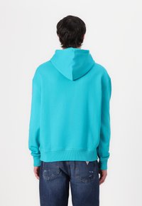 Felpa con cappuccio turchese, dotata di cordoncino, polsini e orlo a coste. Presenta un discreto dettaglio del logo sulla manica sinistra. Indossata sopra un jeans scuro.