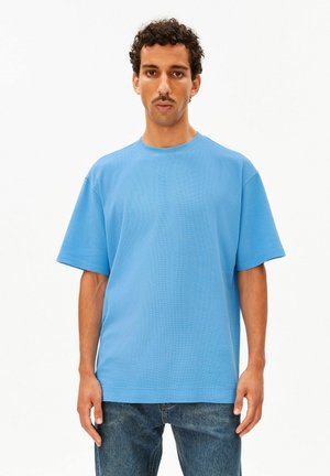 Homme aux cheveux bouclés et à la moustache portant un t-shirt bleu clair uni à manches courtes et un jean bleu, debout devant un fond blanc.