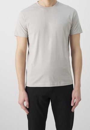 Mand iført en ensfarvet lys grå t-shirt med korte ærmer og sorte bukser, stående med arme afslappet ned langs siderne mod en hvid baggrund.