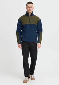Pull en polaire bleu marine et olive, avec un col zippé mi-hauteur, deux poches avant et des panneaux contrastants aux épaules et aux manches.