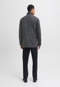 Hall villane mantel kõrge kraega ja tekstuurse viimistlusega, nähtud tagantpoolt. Sobitatud mustade pükste ja elegantsete mustade kingadega.