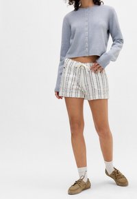 Ljusblå kort kofta med knäppning, kombinerad med vita randiga shorts med resår i midjan och dragsko. Bruna skor.