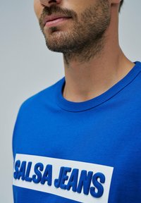 Camiseta de algodón azul con letras texturizadas "SALSA JEANS" en una banda blanca en el pecho. Cuello redondo clásico, superficie lisa.