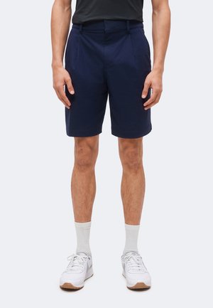 Bărbat stă în picioare purtând pantaloni scurți bleumarin până la genunchi, o cămașă neagră, șosete albe și pantofi sport albi, pe un fundal deschis, simplu.