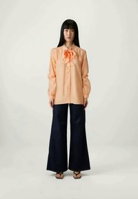 Ανοιχτό πορτοκαλί μπλουζάκι με μακριά μανίκια και φιόγκο στο γιακά, συνδυασμένο με φαρδιά μπλε παντελόνια και μαύρα σαγιονάρες.