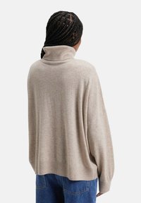 Pull en turtleneck oversize beige en maille douce, avec un ourlet côtelé et des manches amples, porté avec un jean bleu.