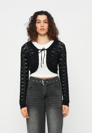Femme aux cheveux bouclés portant un cardigan court noir en maille ouverte noué au col, par-dessus un crop top blanc et un jean taille haute foncé.