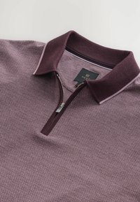 Polo din burgundy cu un model texturat, guler cu fermoar și guler contrastant. Eticheta este vizibilă în interiorul gulerului.