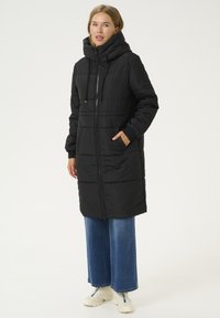 Manteau rembourré noir avec un col haut et une fermeture éclair, arborant un design matelassé et des poches latérales, porté sur un jeans en denim à jambe large.