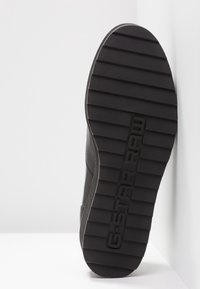 Semelle de chaussure noire avec des rainures horizontales et "G-STAR RAW" embossé au centre, vue d'en dessous sur fond blanc.