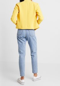Veste jaune cintrée avec une taille ajustée et des manches longues, associée à un jean taille haute bleu clair et des baskets blanches.
