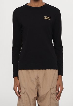 Schwarzes Langarm-T-Shirt aus Baumwolle mit einem kleinen goldenen Logo-Patch auf der Brust und kontrastierenden Seitennähten.