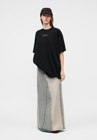 Frau trägt ein schwarzes oversized T-Shirt mit dem Schriftzug "STIEGLITZ", eine schwarze Kappe und einen zweifarbigen langen Jeansrock, steht vor einem weißen Hintergrund.