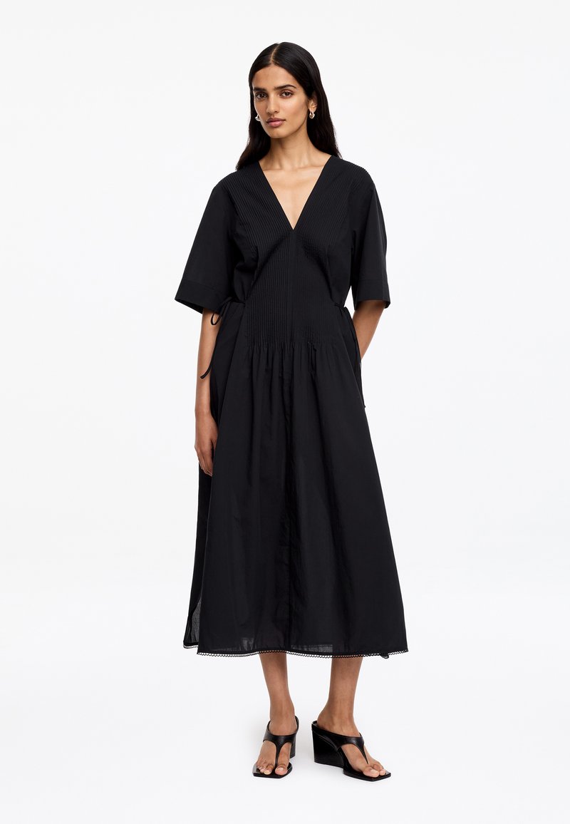 Femme portant une robe midi noire avec un col en V, manches courtes, taille froncée et sandales à talons noires sur fond blanc.