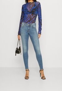 Bodysuit s dlouhým rukávem v modro-černém tygřím vzoru, spárovaný se světle modrými skinny džínami a černými sandály na podpatku, držící malou černou kabelku.