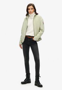 Giacca in pile verde chiaro, con zip, indossata sopra un maglione bianco a treccia e jeans skinny neri. Stivali neri con lacci completano l'outfit.