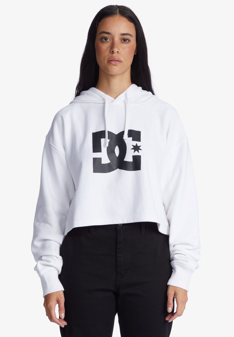Weißer, cropped Hoodie mit schwarzem DC-Logo und Stern-Design. Verfügt über eine Kapuze mit Kordelzug und lange Ärmel, kombiniert mit schwarzen Hosen.