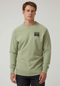 Hombre vistiendo un sudadera verde claro con el texto "DREAM BIG ALTONADOCK" en el pecho, de pie frente a un fondo claro y liso.