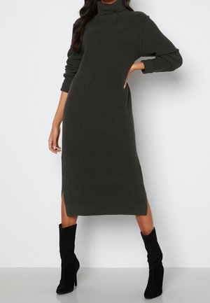 Kobieta ubrana w ciemnozieloną prążkowaną, dzianinową sukienkę midi z golfem, długimi rękawami i bocznymi rozcięciami, w połączeniu z czarnymi botkami na obcasie.