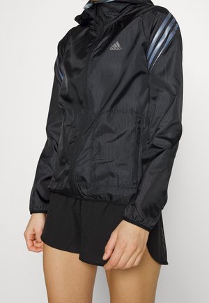 Personne portant une veste zippée Adidas noire avec des bandes réfléchissantes sur les manches et un short de sport noir sur fond blanc uni.