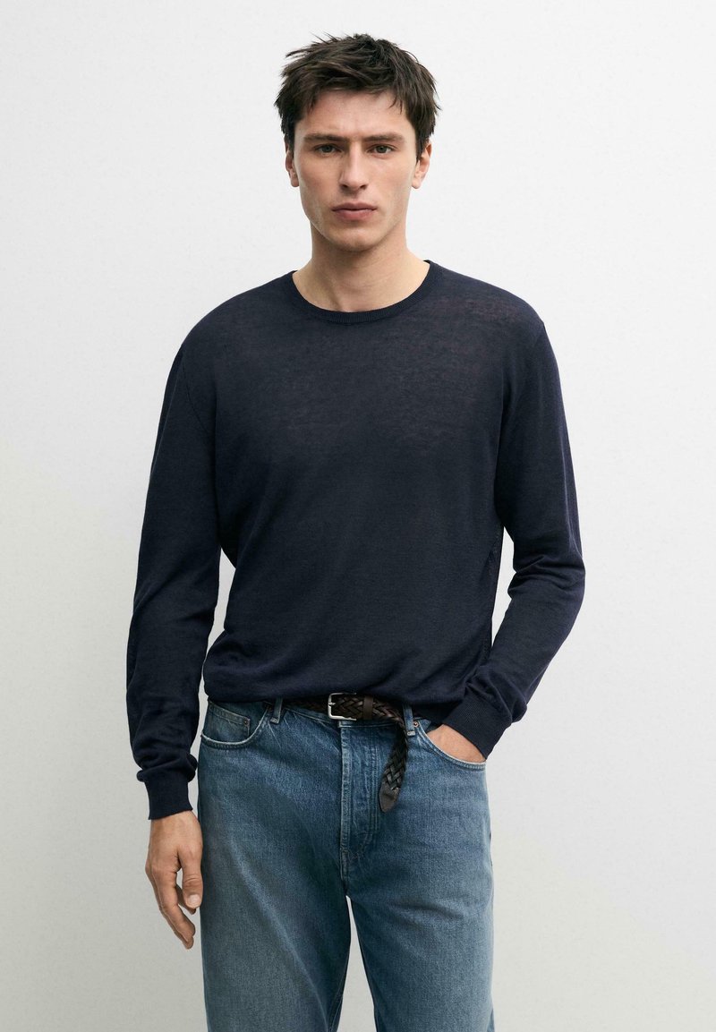 Uomo con maglione blu scuro a maniche lunghe e jeans blu con cintura intrecciata, in piedi davanti a uno sfondo chiaro e uniforme.