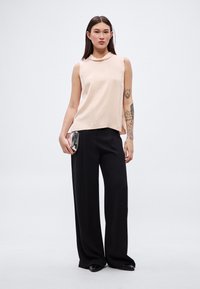 Saténový tielkový top v jemnej broskyňovej farbe v kombinácii s čiernymi širokými nohavicami, ktoré majú hladký povrch a jemný závesný efekt. Model drží malú striebornú kabelku.