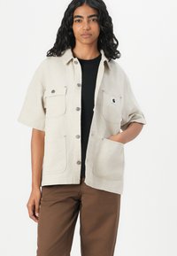 Beige short-sleeve shirt jacket with two front pockets, button closure, and a logo patch, made from textured cotton fabric.
