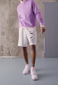 Fioletowa bluza na zamek z różowym kołnierzem, białe spodenki z wieloma czarnymi logo Nike oraz białe trampki z kolorowymi skarpetkami w paski.