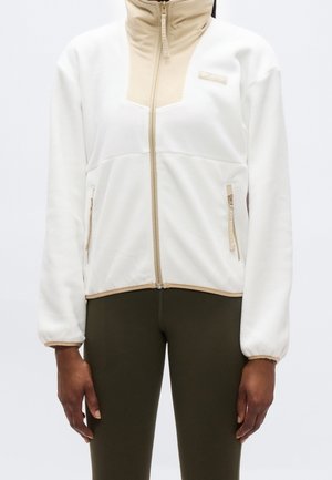 Weiße Fleecejacke mit beigen Reißverschluss- und Kragen-Details, getragen zu dunkelolivfarbenen Leggings, Ansicht von den Schultern bis zur Hüfte.