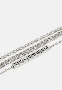 Pulseira prateada de múltiplas camadas com três correntes: uma com strass, uma corrente grossa e uma com blocos de letras que soletram "uncommon."