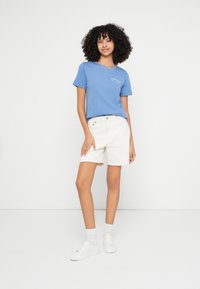 Kék pamut rövid ujjú póló fehér logóval, fehér magas derekú rövidnadrággal párosítva. A modell fehér tornacipőt és zoknit visel.