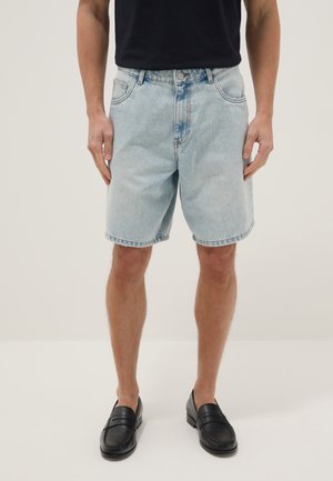 Mann trägt hellblaue Jeansshorts, schwarzes T-Shirt und schwarze Loafers, steht mit entspannten Händen seitlich an weißem Hintergrund.