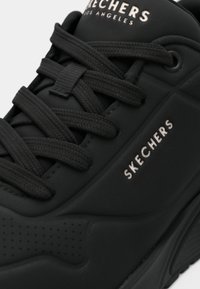 Schwarzer Skechers-Sneaker mit strukturiertem Gummi-Obermaterial, flachen schwarzen Schnürsenkeln, silbernem Branding und glatter Sohle.