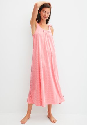 Jonge vrouw die blootsvoets staat, gekleed in een losse, mouwloze midi-jurk met rood-witte strepen en knoopbandjes, op een witte achtergrond.