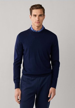 Mann mit kurzen braunen Haaren, der einen dunkelblauen Pullover über einem blauen Hemd mit Kragen und passende dunkelblaue Hose trägt, steht vor einem einfarbigen Hintergrund.
