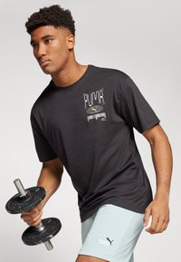 Camiseta de algodón negra con un diseño gráfico en el lado izquierdo del pecho, combinada con shorts de color azul claro, sosteniendo una barra con pesas negras.