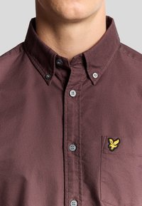 Camicia button-up color borgogna con colletto strutturato, una tasca sul petto con un logo giallo ricamato e bottoni grigi.