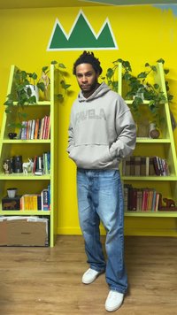 Sweat à capuche gris avec "FAVELA" imprimé en lettres majuscules, associé à un jean bleu délavé et des baskets blanches. Étagères vertes et mur jaune en arrière-plan.