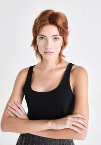 Svart ermeløs tanktop med rund hals, laget av glatt stoff. Modellen har kort, teksturert rødt hår og armene krysset.