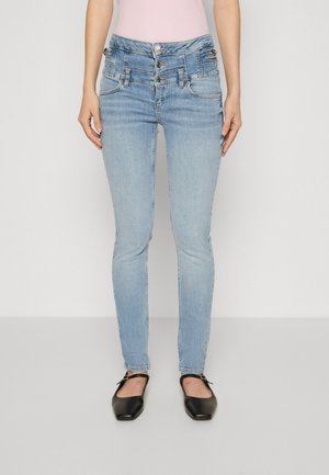 Donna che indossa jeans skinny a doppia vita azzurri chiaro e ballerine nere a punta, in piedi davanti a uno sfondo chiaro uniforme.