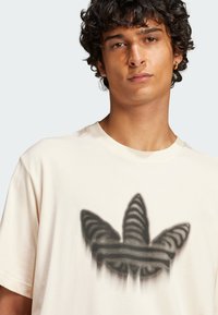 T-shirt crema in cotone con una grande grafica nera sbiadita di un logo a tre foglie sulla parte anteriore. Maniche corte e collo rotondo.