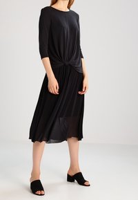 Svart klänning med en knuten detalj vid midjan, knälång, långärmad, flödande kjol, tillsammans med svarta öppna-toe mules.