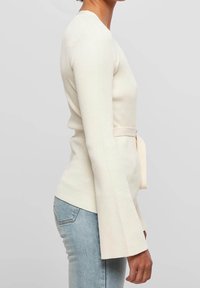 Ljusgräddig, ribbad långärmad topp med en åtsittande silhuett, utsvängda ärmar och en knuten midjedetalj. Matchad med ljusblå denimjeans.