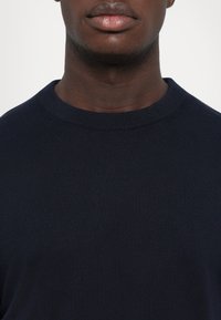 Lähivõte inimese alumisest näost ja kaelast, kes kannab tavalist tumesinist ümmarguse kaelusega särk, lihtsa tausta ees.