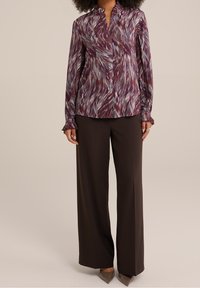 Långärmad blus i ett lila, flerfärgat abstrakt mönster, med volangkrage och manschetter, i kombination med skräddarsydda mörkbruna byxor.