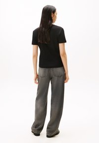 Hosszú sötét hajú nő fekete rövid ujjú pólóban és laza szürke farmernadrágban, háttal áll egy egyszerű fehér háttér előtt.