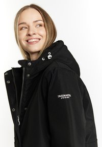Sort hættetrøje med lynlås, trykknaplukning og broderet logo. Har en blød tekstur og justerbar snøre i hætten.
